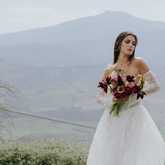 A woman in a white wedding dress holds a bouquet of flowers, standing outdoors with mountains and greenery in the background.