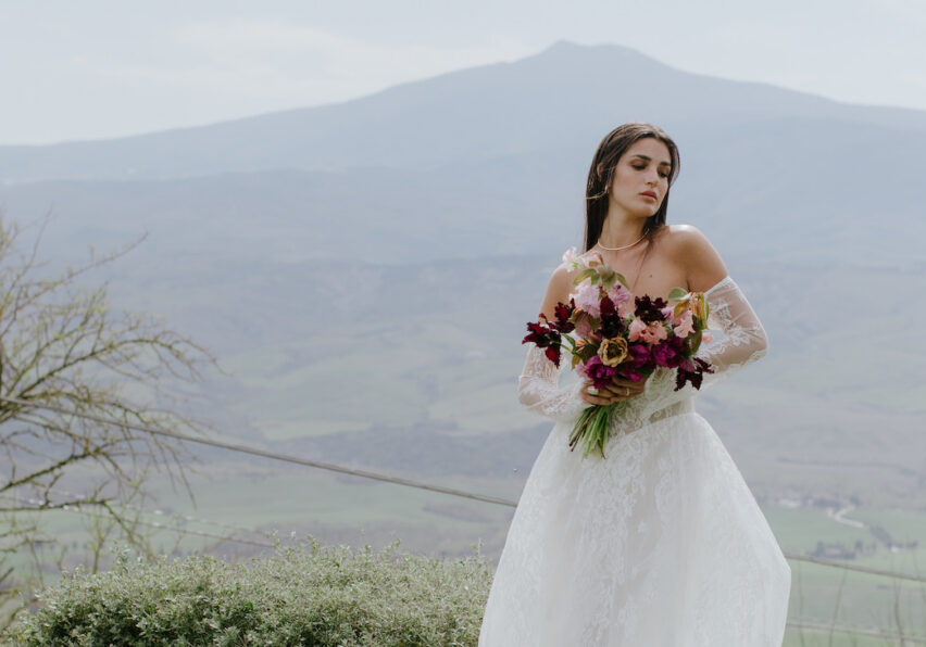 A woman in a white wedding dress holds a bouquet of flowers, standing outdoors with mountains and greenery in the background.