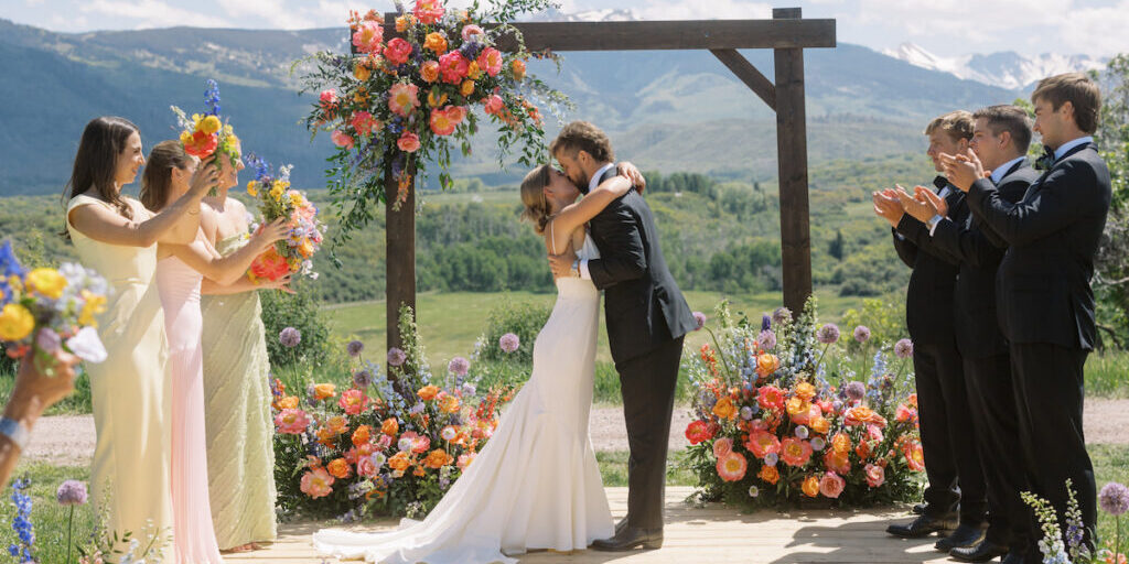 A bride and groom embrace and kiss on an outdoor wooden platform decorated with colorful flowers, surrounded by applauding wedding party members, with mountains and greenery in the background.