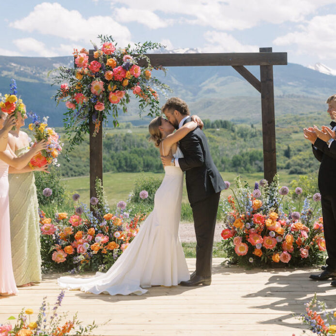 A bride and groom embrace and kiss on an outdoor wooden platform decorated with colorful flowers, surrounded by applauding wedding party members, with mountains and greenery in the background.