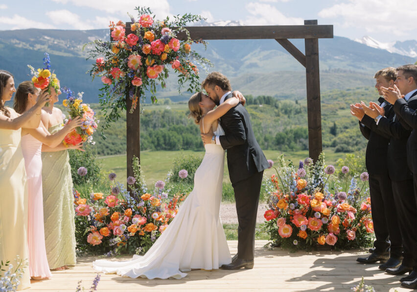A bride and groom embrace and kiss on an outdoor wooden platform decorated with colorful flowers, surrounded by applauding wedding party members, with mountains and greenery in the background.
