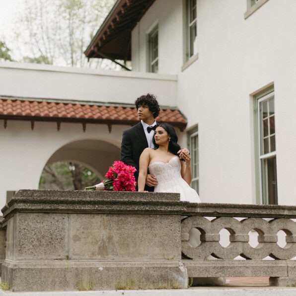 A bride and groom stand together outside by a stone railing, the bride holding a bouquet of bright pink flowers.