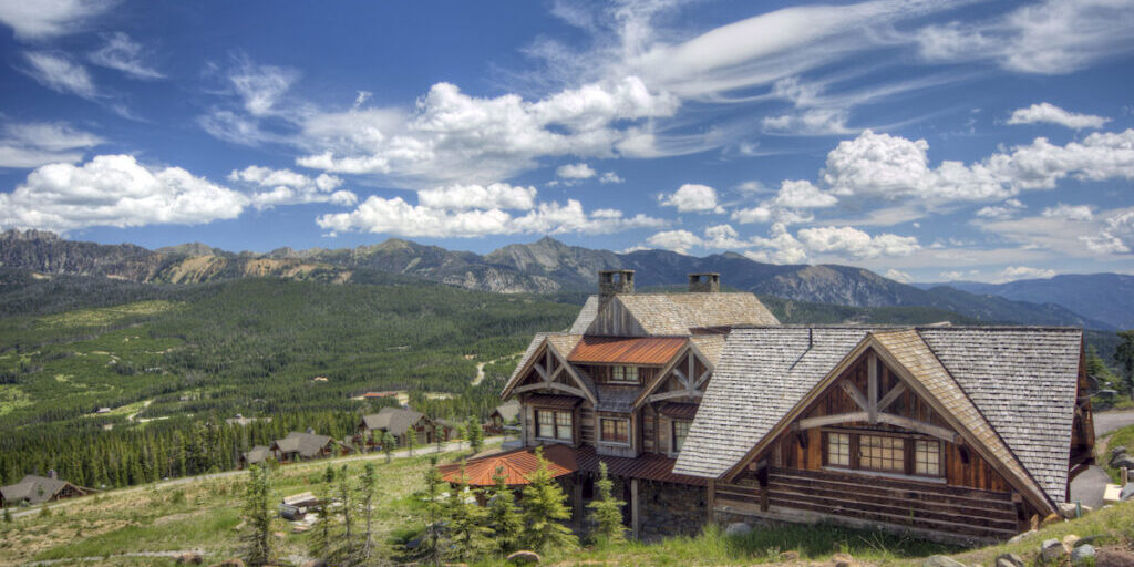 A rustic wooden lodge sits on a hillside with mountains, forests, and scattered houses visible under a partly cloudy blue sky.