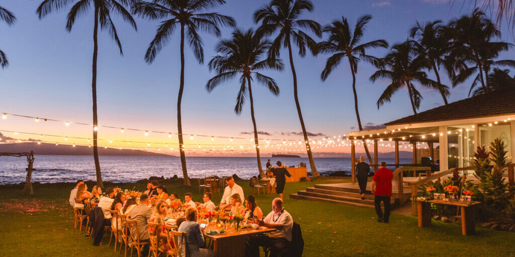 People dining outdoors at long tables under string lights near the ocean at sunset, with palm trees and a wooden structure in the background.