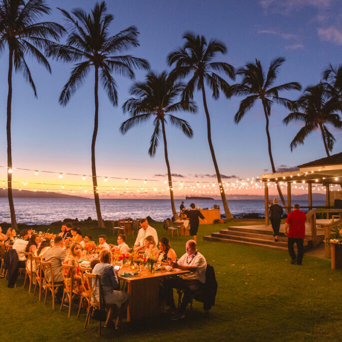 People dining outdoors at long tables under string lights near the ocean at sunset, with palm trees and a wooden structure in the background.