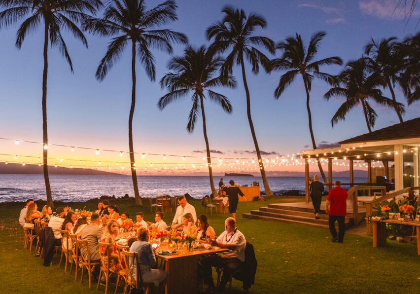 People dining outdoors at long tables under string lights near the ocean at sunset, with palm trees and a wooden structure in the background.