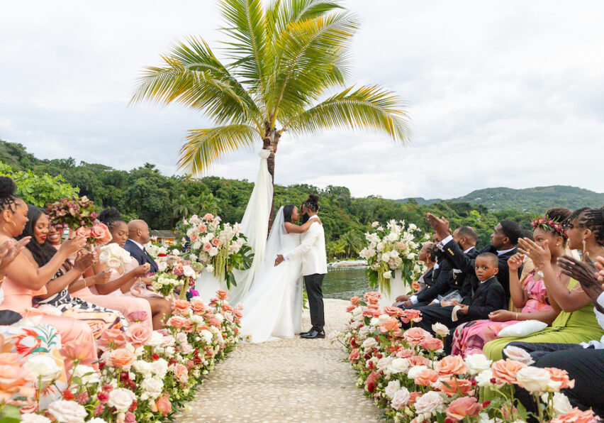 A couple kisses at an outdoor wedding ceremony, surrounded by guests seated along an aisle lined with flowers, with a palm tree and greenery in the background.