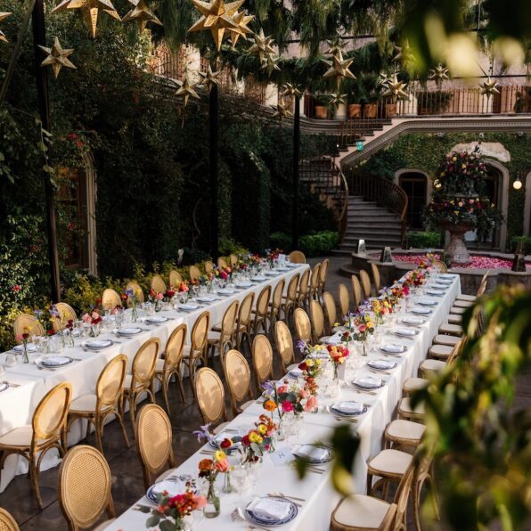 Long banquet tables with white tablecloths and gold chairs are set for an outdoor event, surrounded by greenery and star-shaped hanging decorations.
