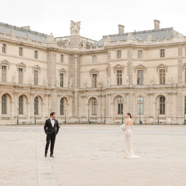 A man in a tuxedo and a woman in a white dress stand apart in an empty courtyard in front of a historic building. Featured image for Your Guide to Iconic Paris Wedding Photos