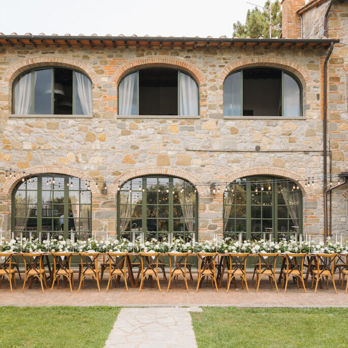 Long dining table set for an outdoor event, with white flowers and candles, placed in front of a rustic stone building.