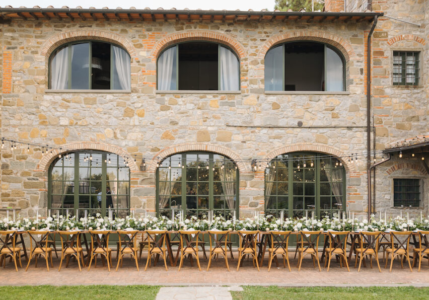 Long dining table set for an outdoor event, with white flowers and candles, placed in front of a rustic stone building.