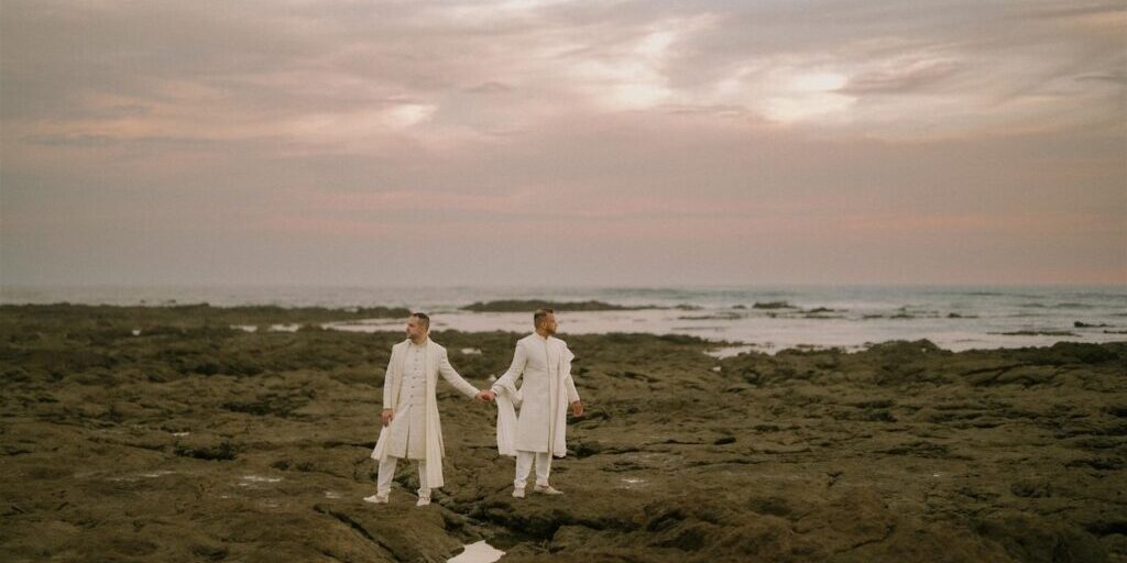 Two people dressed in white stand holding hands on a rocky shoreline at sunset, with the ocean and a cloudy sky in the background.