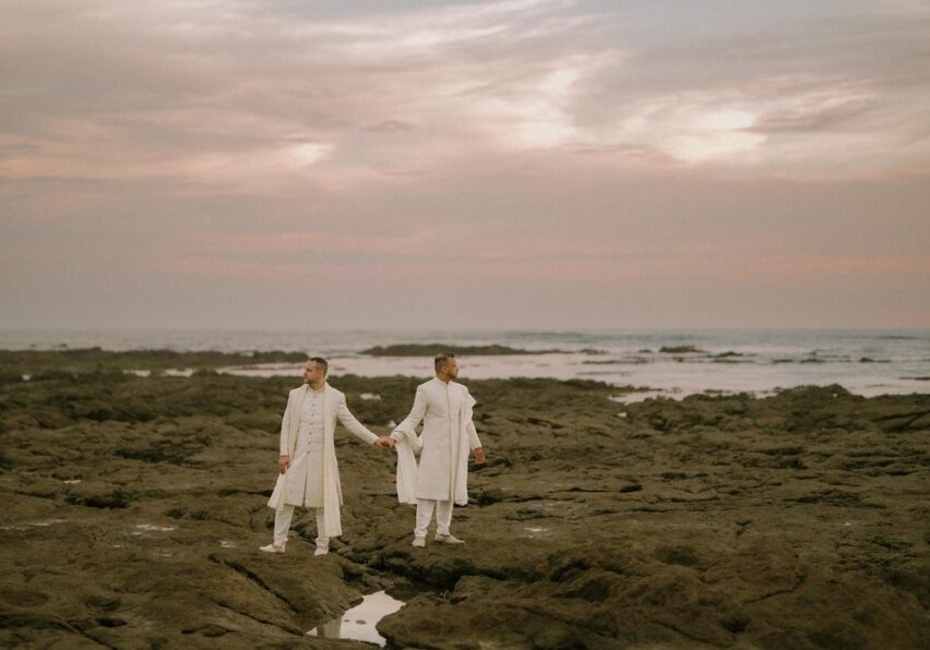 Two people dressed in white stand holding hands on a rocky shoreline at sunset, with the ocean and a cloudy sky in the background.