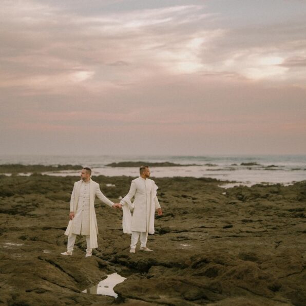 Two people dressed in white stand holding hands on a rocky shoreline at sunset, with the ocean and a cloudy sky in the background.
