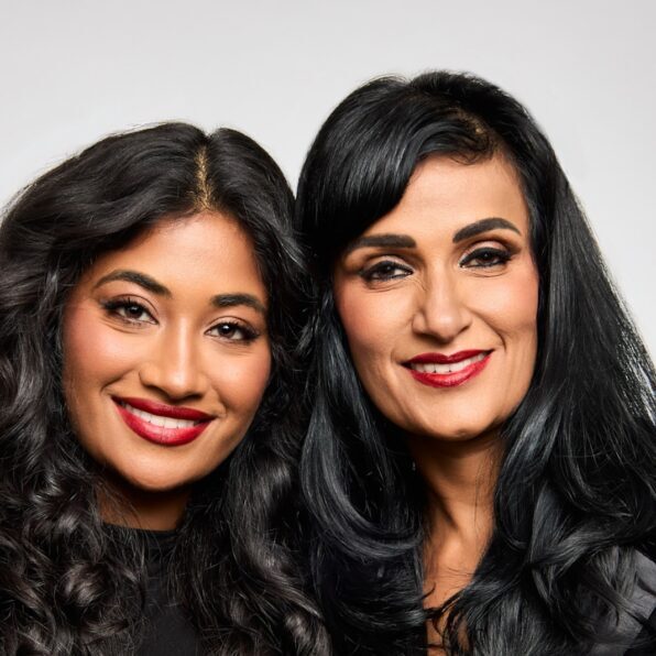 Two women with long black hair and red lipstick smile, standing close together in front of a plain light background.