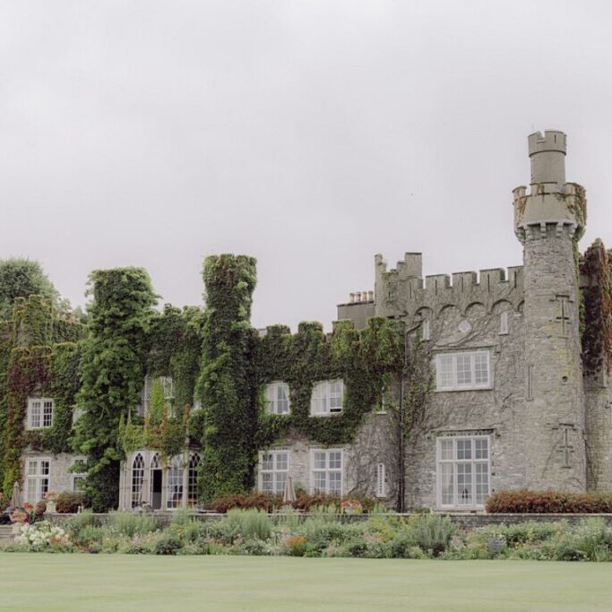 A large stone castle covered in ivy with multiple towers and windows, surrounded by a neatly maintained garden and expansive lawn. The sky above is overcast.