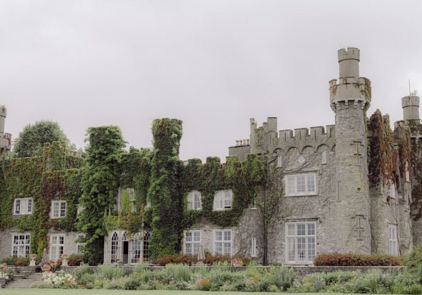 A large stone castle covered in ivy with multiple towers and windows, surrounded by a neatly maintained garden and expansive lawn. The sky above is overcast.