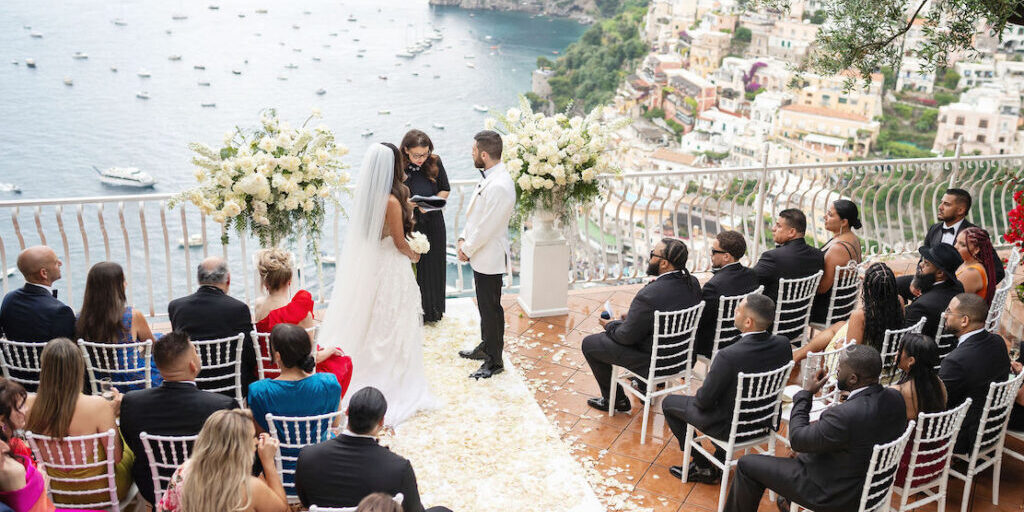 A bride and groom stand before an officiant at an outdoor wedding ceremony overlooking the sea, with guests seated in rows on a terrace.