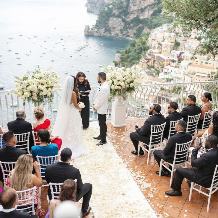 A bride and groom stand before an officiant at an outdoor wedding ceremony overlooking the sea, with guests seated in rows on a terrace.