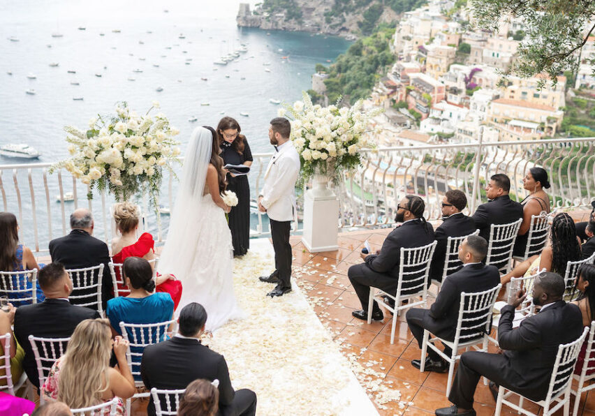 A bride and groom stand before an officiant at an outdoor wedding ceremony overlooking the sea, with guests seated in rows on a terrace.