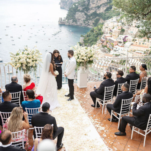 A bride and groom stand before an officiant at an outdoor wedding ceremony overlooking the sea, with guests seated in rows on a terrace.