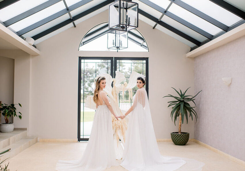 Two women in white gowns stand indoors, holding hands and facing the camera, with a large window, sculpture, and potted plant behind them.