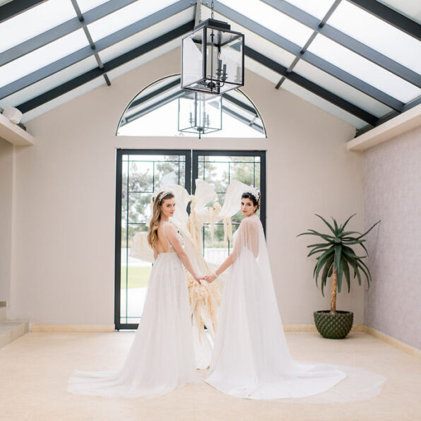Two women in white gowns stand indoors, holding hands and facing the camera, with a large window, sculpture, and potted plant behind them.