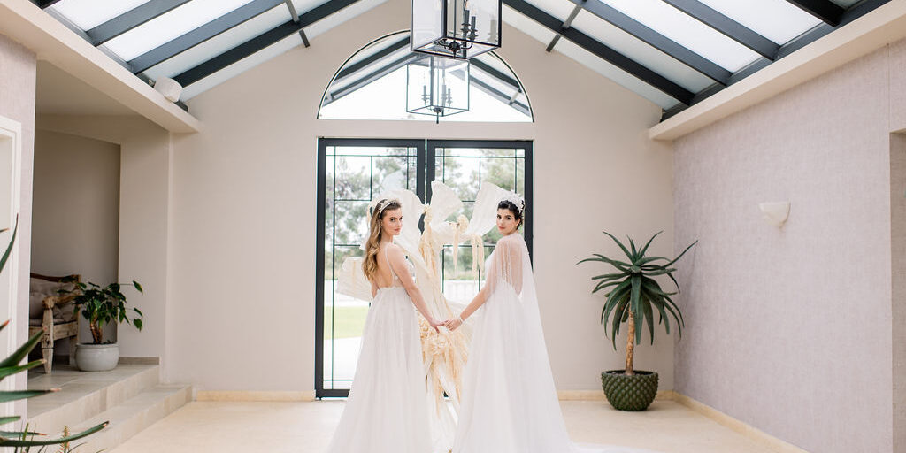 Two women in white gowns stand indoors, holding hands and facing the camera, with a large window, sculpture, and potted plant behind them.