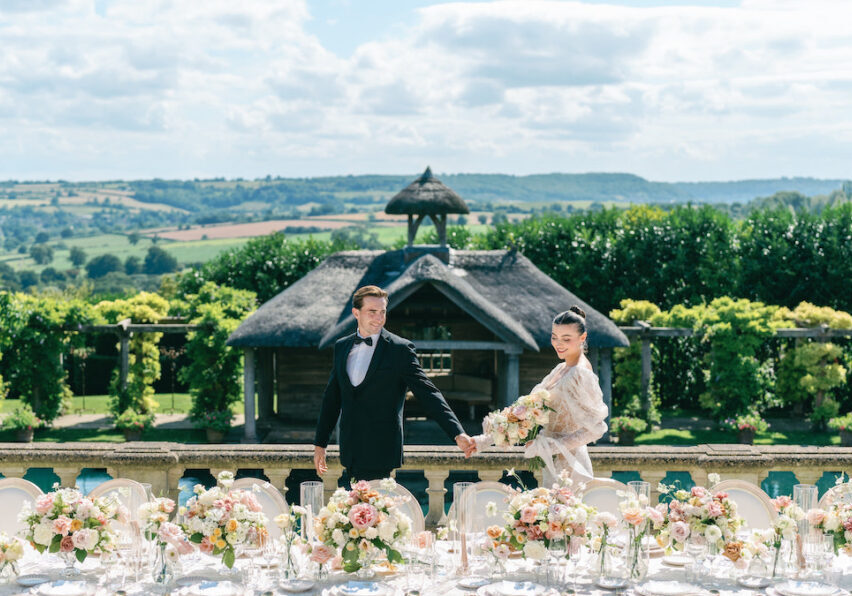 A couple in formal attire poses in a manicured garden with a pool and wooden pavilion.