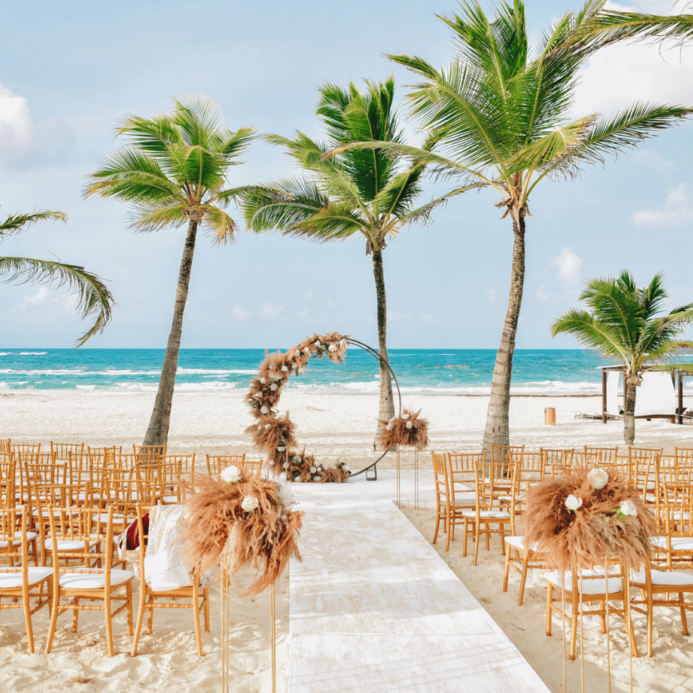 Beach wedding setup with wooden chairs facing an ocean backdrop, palm trees, and a decorated circular arch under a clear sky.