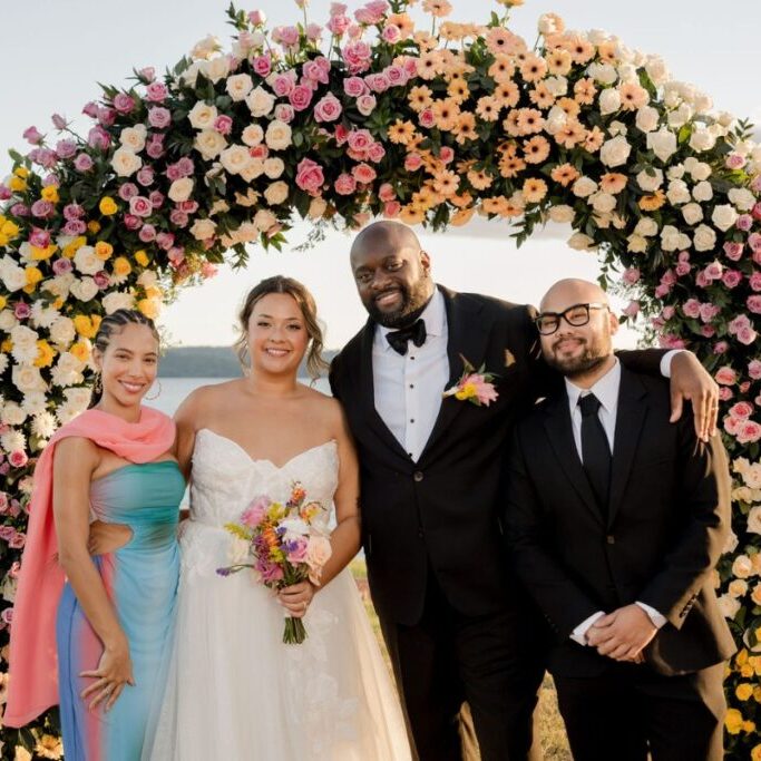 Four people stand smiling under a large floral arch; one in a wedding dress, one in a tuxedo, and two in formal attire.