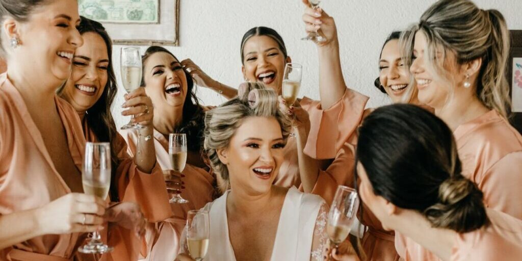 A bride with hair rollers in her hair sits surrounded by smiling bridesmaids holding glasses of champagne.