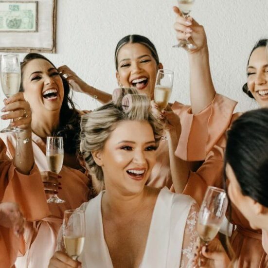 A bride with hair rollers in her hair sits surrounded by smiling bridesmaids holding glasses of champagne.