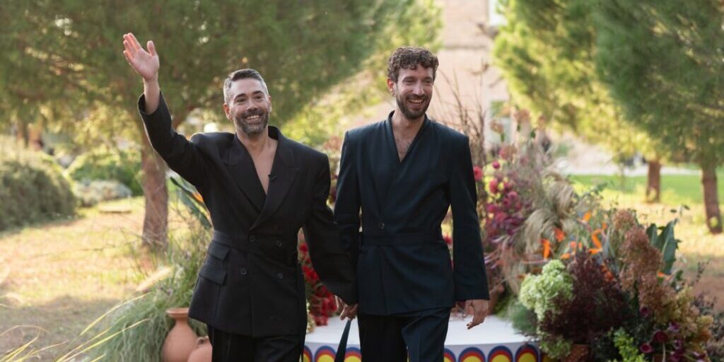 Two men in black suits hold hands and smile while walking outdoors, with one waving. Decorative plants and a small stage are in the background.