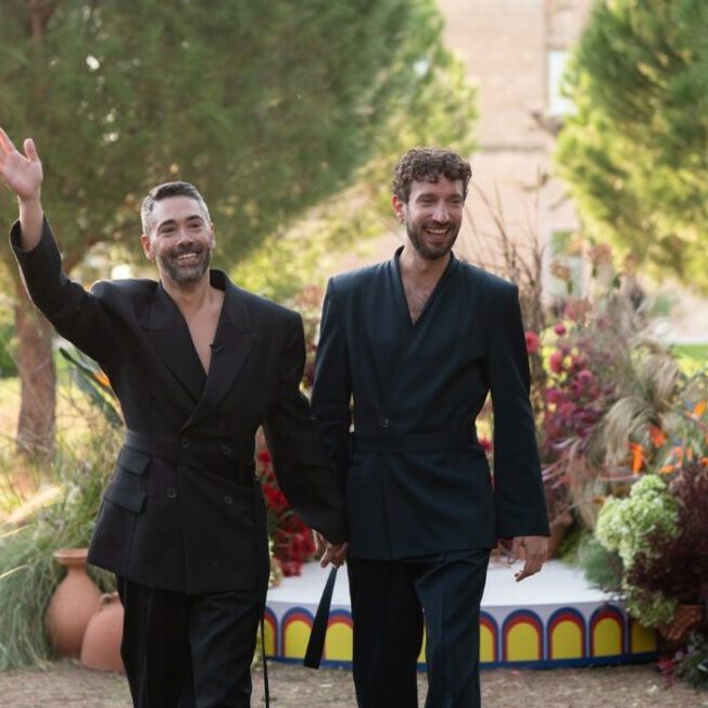 Two men in black suits hold hands and smile while walking outdoors, with one waving. Decorative plants and a small stage are in the background.