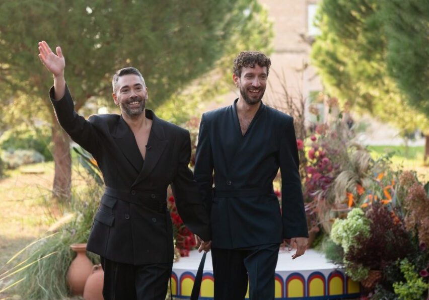 Two men in black suits hold hands and smile while walking outdoors, with one waving. Decorative plants and a small stage are in the background.