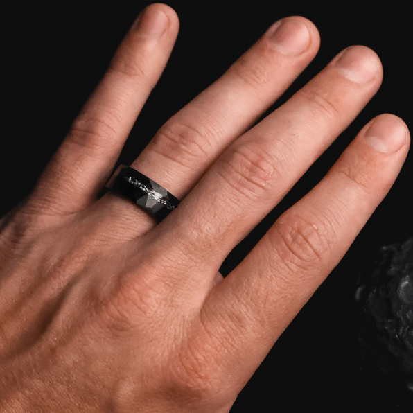 A hand wearing a black and silver wedding ring with a textured band rests on a smooth surface.