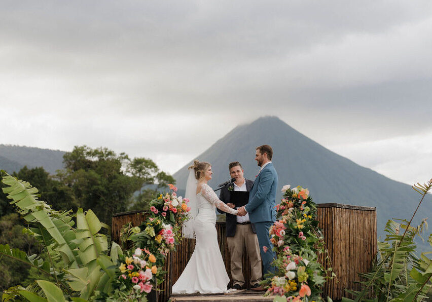 A bride and groom stand with an officiant on a flower-decorated platform outdoors, with a mountain visible in the background under a cloudy sky.