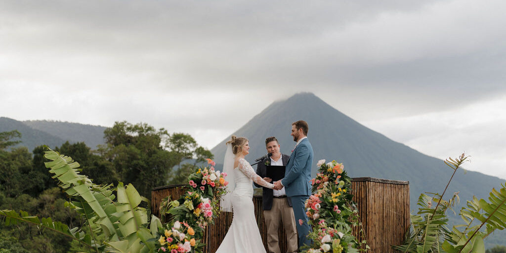 A bride and groom stand with an officiant on a flower-decorated platform outdoors, with a mountain visible in the background under a cloudy sky.