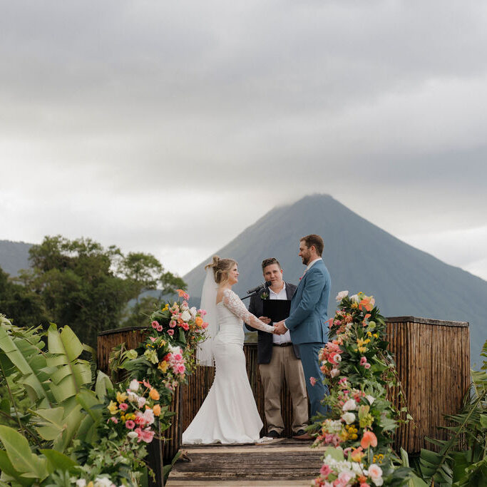 A bride and groom stand with an officiant on a flower-decorated platform outdoors, with a mountain visible in the background under a cloudy sky.