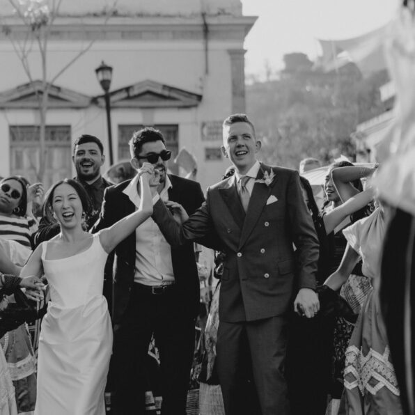 A black and white image of a wedding procession with a joyful bride and groom holding hands, surrounded by smiling, formally dressed guests and performers in traditional attire.