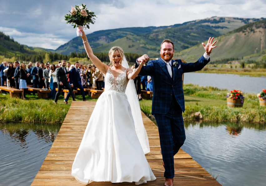 A bride and groom walk hand in hand down a wooden walkway over water, with a cheering crowd and mountainous landscape in the background.