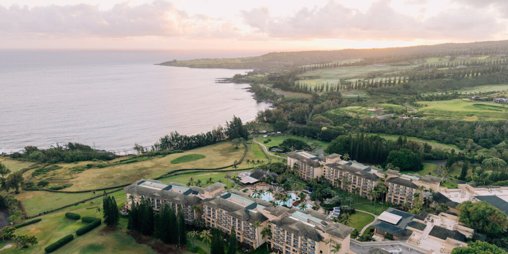 Aerial view of a large coastal resort surrounded by golf courses and greenery, with the ocean and a hilly landscape in the background at sunset.