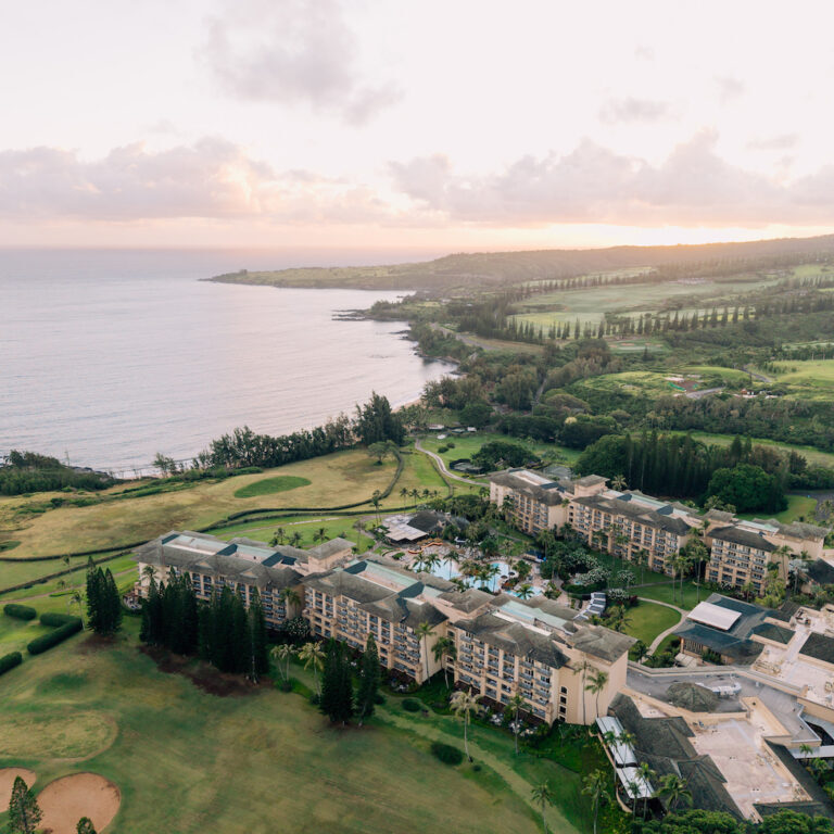 Aerial view of a large coastal resort surrounded by golf courses and greenery, with the ocean and a hilly landscape in the background at sunset.