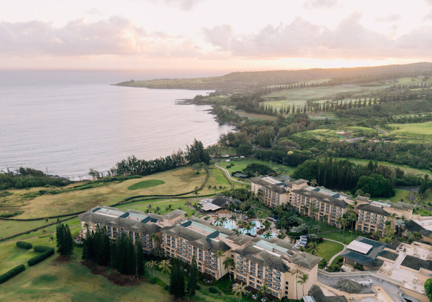 Aerial view of a large coastal resort surrounded by golf courses and greenery, with the ocean and a hilly landscape in the background at sunset.