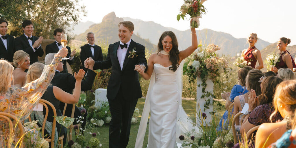 A bride and groom walk hand-in-hand down an outdoor wedding aisle, holding hands and smiling toward guests.