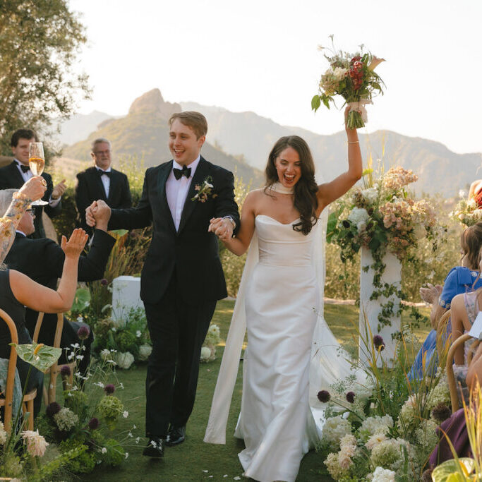 A bride and groom walk hand-in-hand down an outdoor wedding aisle, holding hands and smiling toward guests.