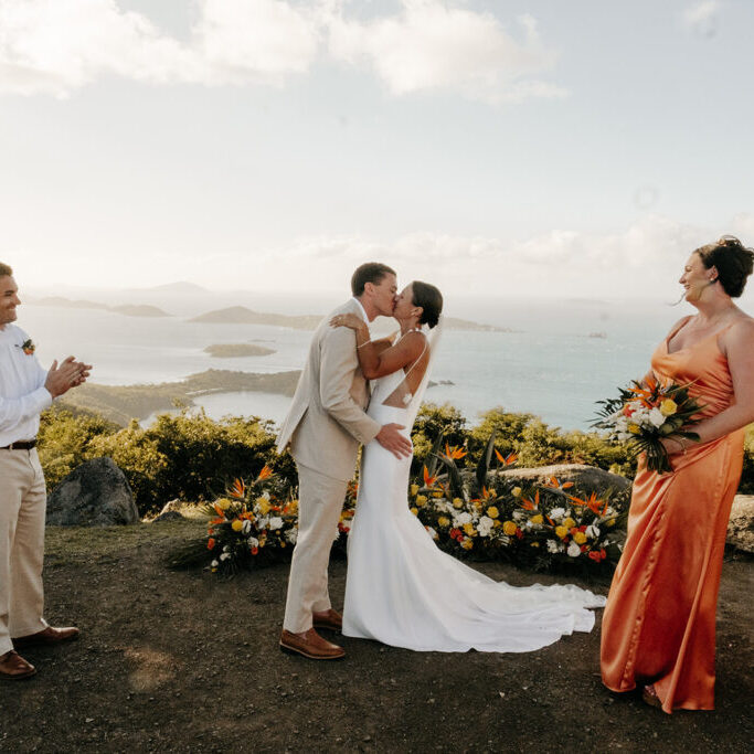 A bride and groom kiss outdoors at their wedding ceremony, with two attendants standing nearby and scenic ocean views in the background.