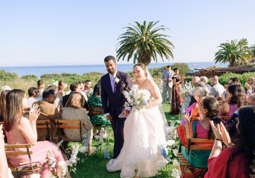 A newlywed couple walks down an outdoor aisle, surrounded by seated guests. The bride is wearing a white wedding dress and holding a bouquet, while the groom is in a dark suit. The ocean and palm trees can be seen in the background under a clear blue sky.