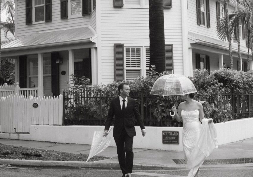 A man in a suit and a woman in a wedding dress cross a street holding transparent umbrellas in front of the Hemingway House on a rainy day in Key West, Florida.